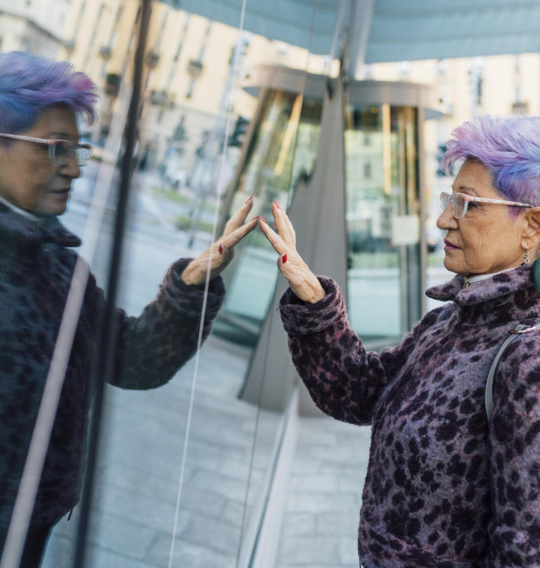 Elderly woman with stylish purple hair admires her reflection in a modern cityscape.